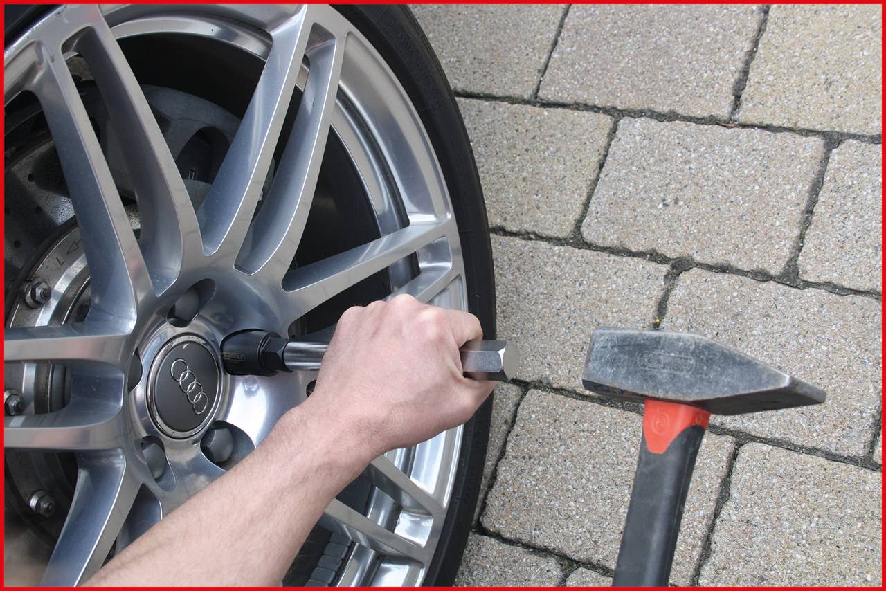 A person grasps the wheel of a vehicle with their hand, using a tool to tighten screws. Close-up of the alloy rim and cobblestone surface.