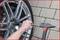 A person grasps the wheel of a vehicle with their hand, using a tool to tighten screws. Close-up of the alloy rim and cobblestone surface.