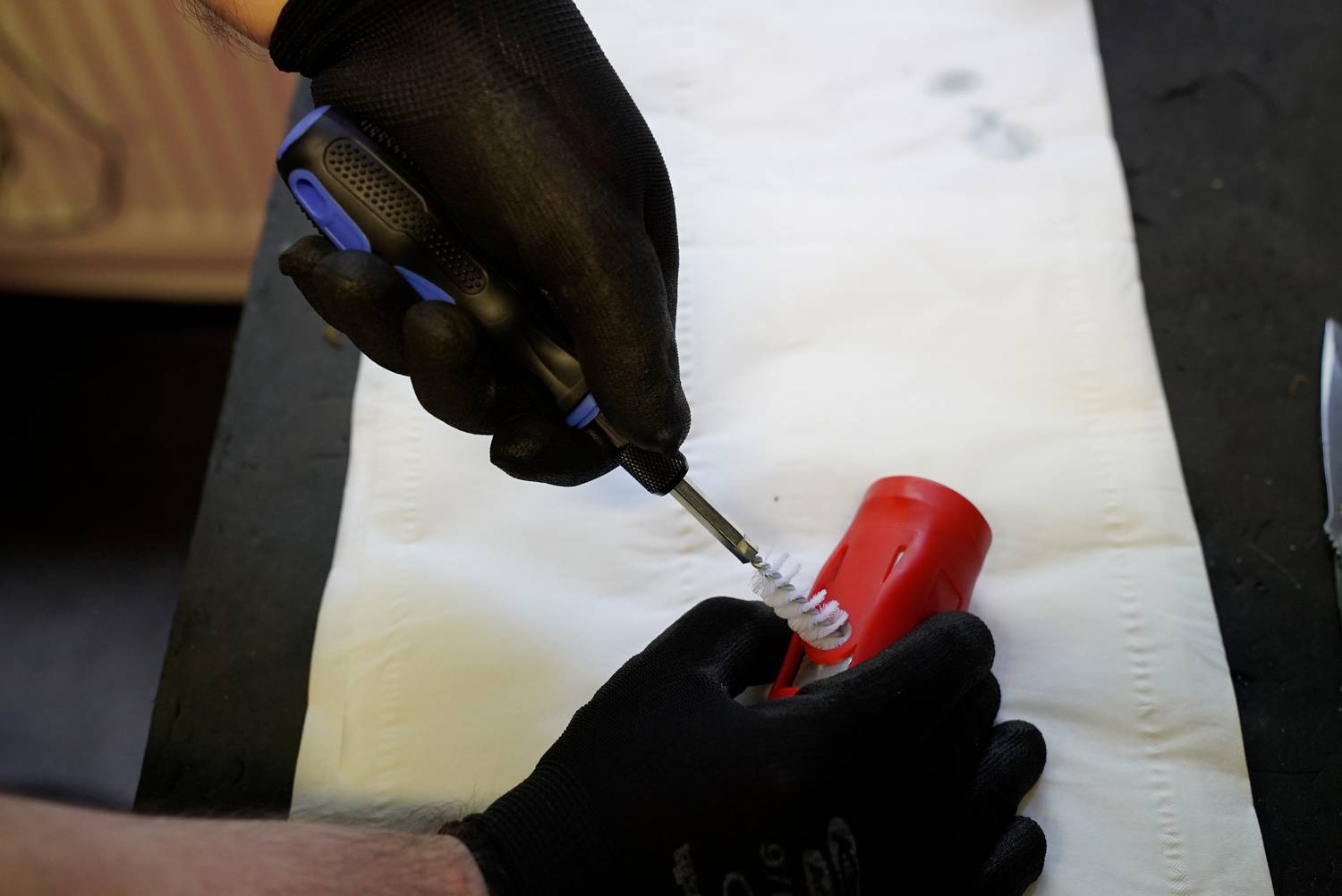 A hand wearing black gloves cleans a red metal part with a brush on a white background.