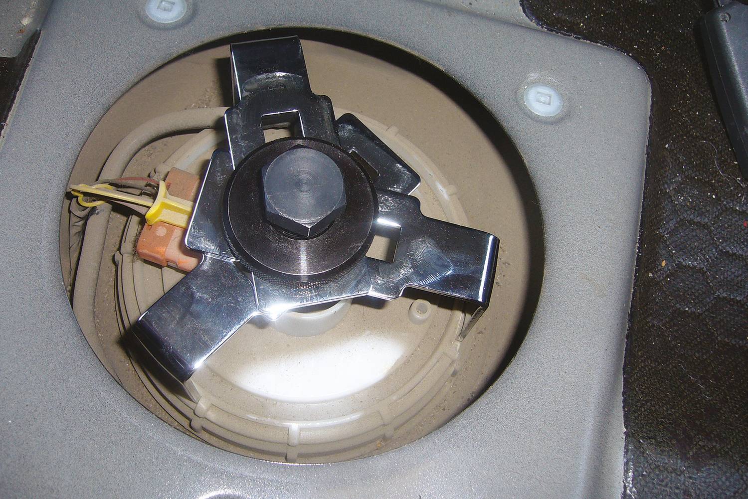 A car fuel filler cap from the inside, with a silver locking mechanism surrounded by cables. The area is dusty.