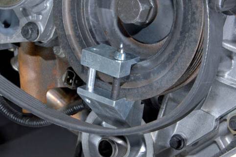 Close-up of a V-ribbed belt wound around a tensioner pulley in an engine. A clamp holds the belt in place.