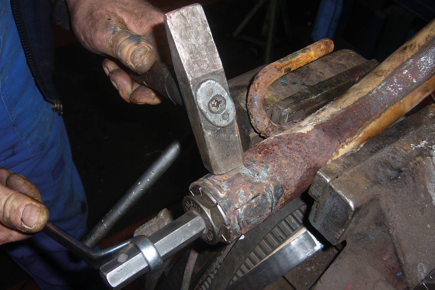 A tradesman is using a spanner and a hammer to work on a rusty metal pipe that is clamped in a vice.