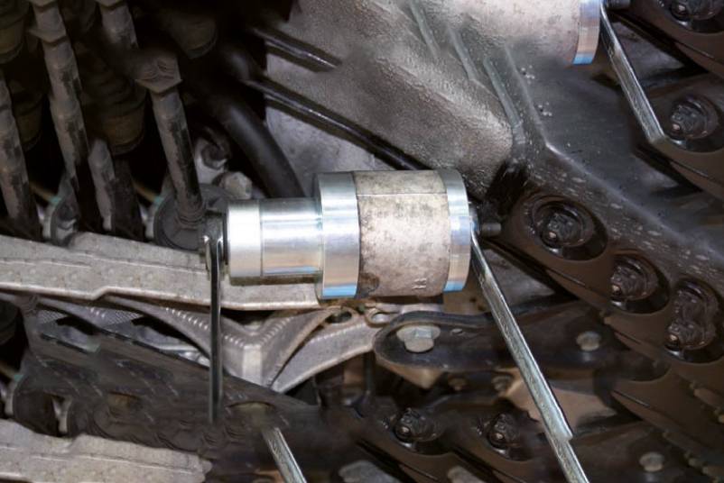A metal tool is attached to a chain drive in an engine compartment. It appears to be used for adjustment or maintenance.