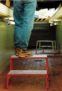 A person wearing jeans and leather boots is standing on a red two-step stepladder in a narrow corridor.