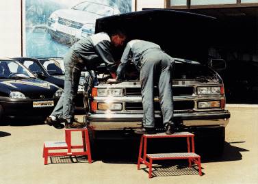 Two mechanics are repairing the engine of a pick-up truck in a workshop. They are standing on red steps to gain better access.