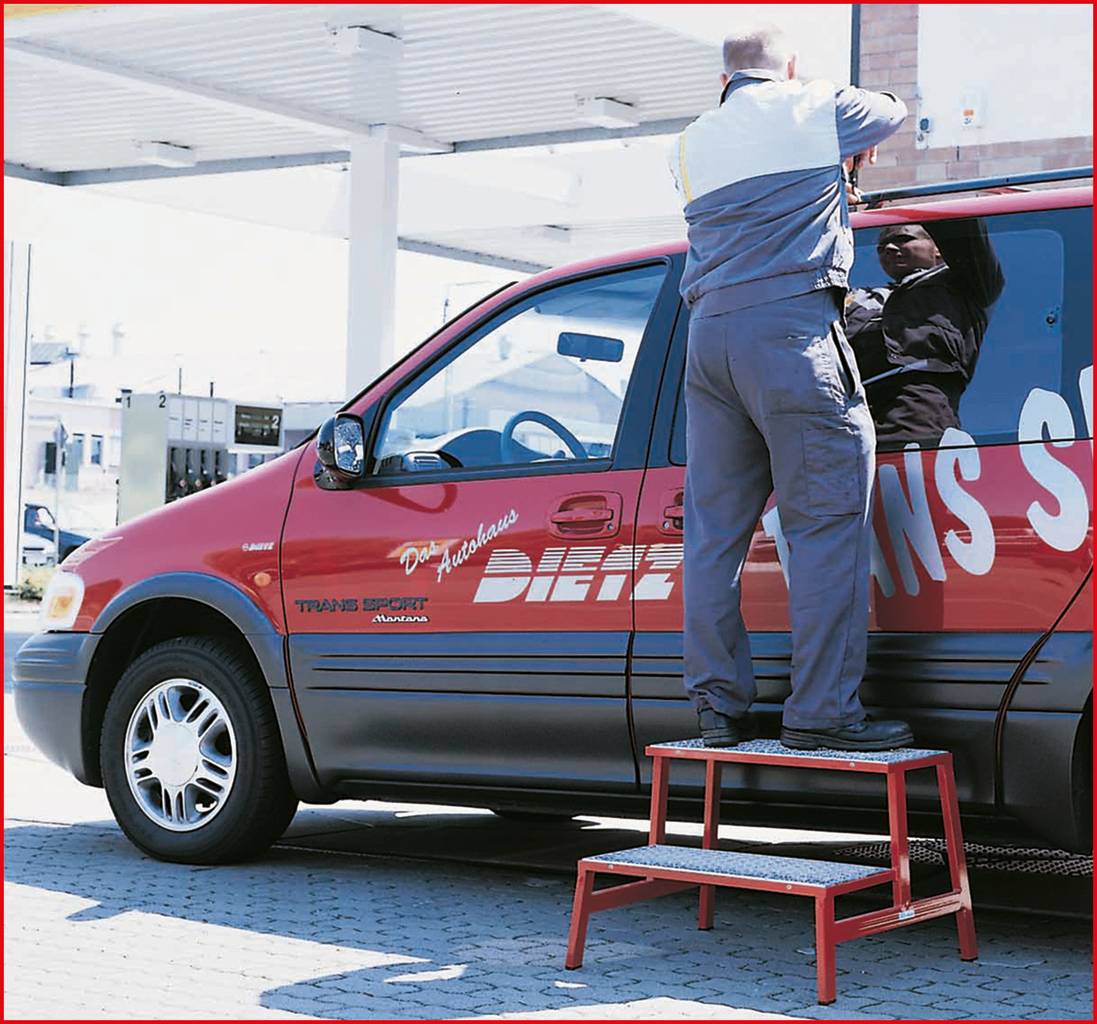 A man in workwear is installing a mirror on a red van at a petrol station. A ladder is standing next to the vehicle.