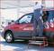 A man in workwear is installing a mirror on a red van at a petrol station. A ladder is standing next to the vehicle.