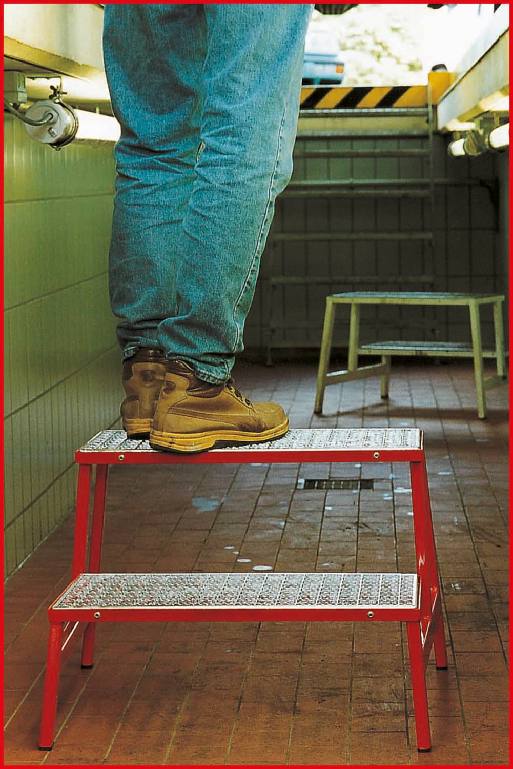 A person is standing on a red two-step folding ladder in a tiled room, wearing jeans and brown work boots.