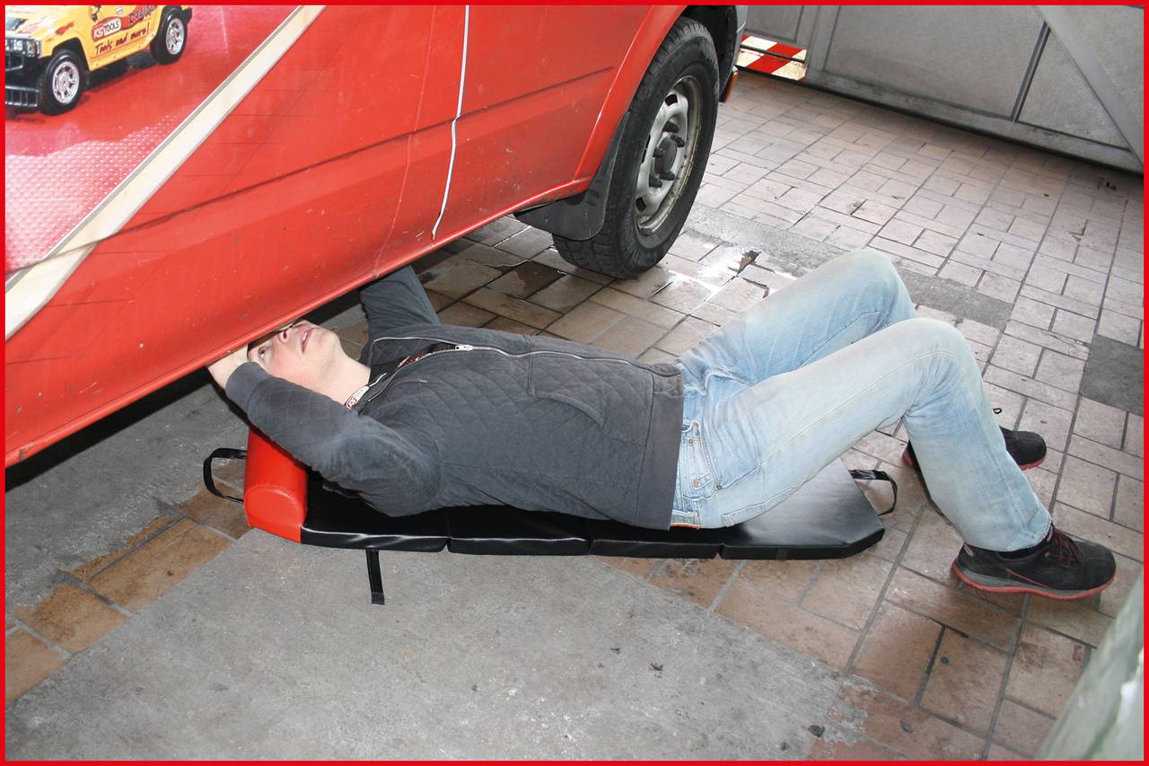 A person is lying on a trolley underneath a red delivery van, apparently carrying out repair work in a garage.