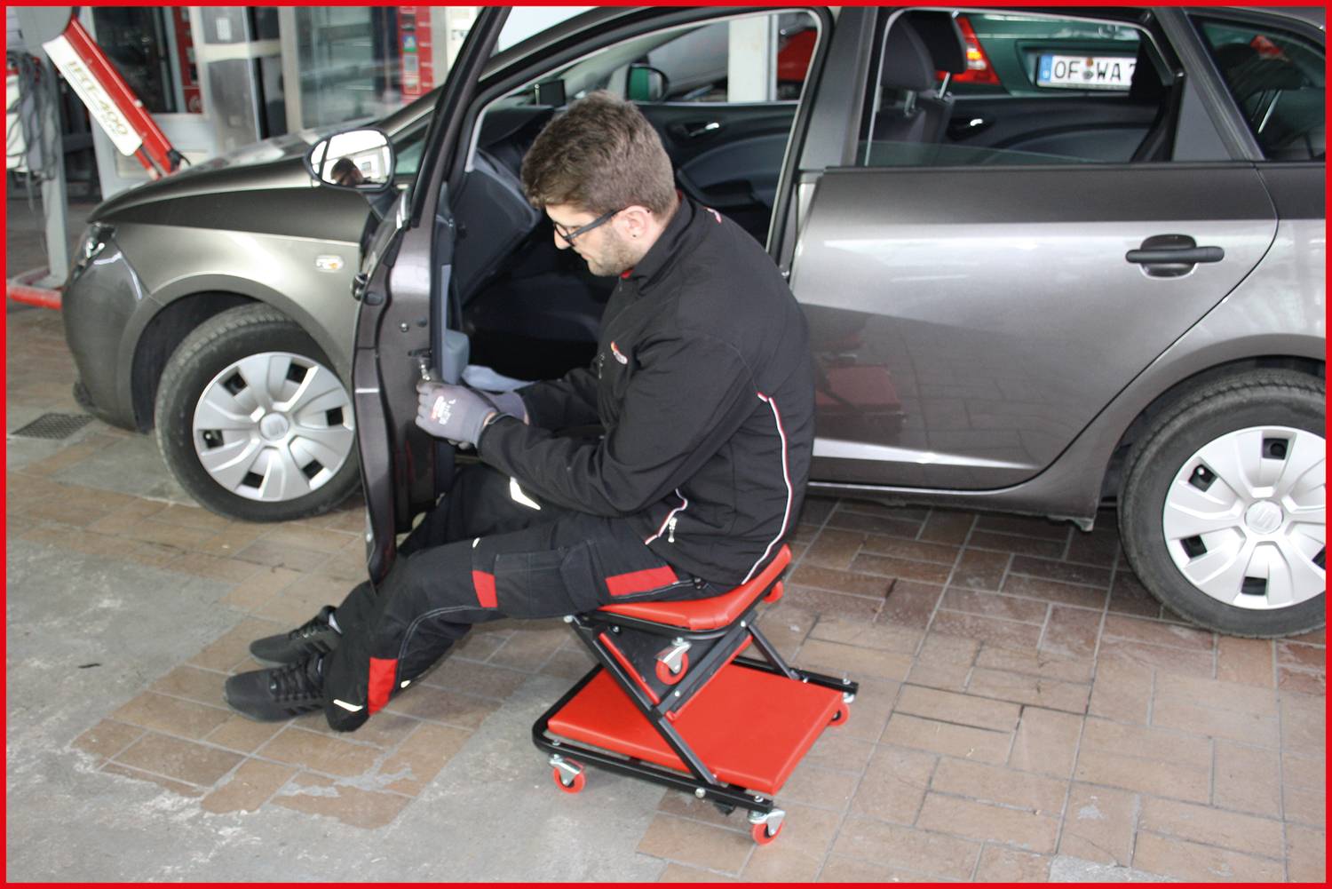 A mechanic wearing black workwear sits on a workshop stool and works on the open car door of a grey saloon car.