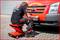 A mechanic sits on a wheeled stool and works on the front bumper of a red Ford van on a paved floor.
