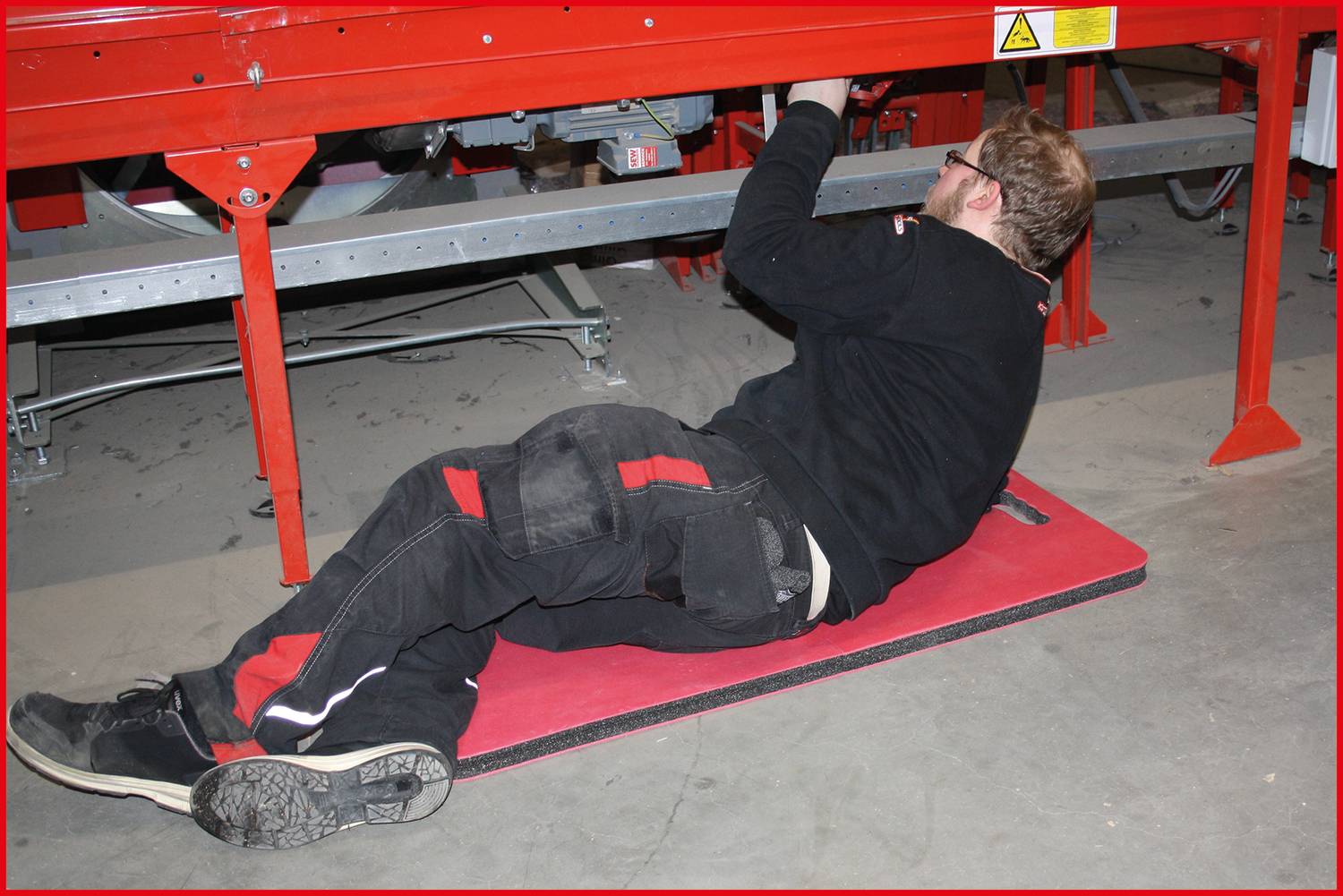 A technician is lying underneath a red machine, working on it. He is wearing work clothing and safety glasses.
