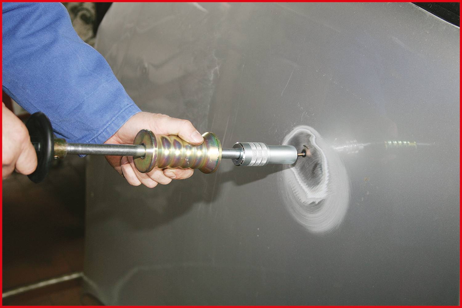 A person is repairing a dent in a car door with specialist tools, using a pulling hammer to smooth out the metal.