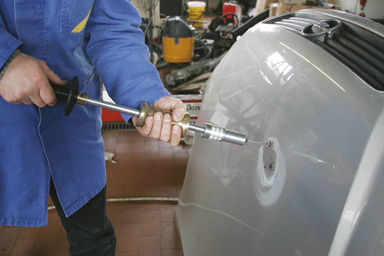 A person in blue workwear is repairing a large metal surface with a tool. Workshop environment in the background.