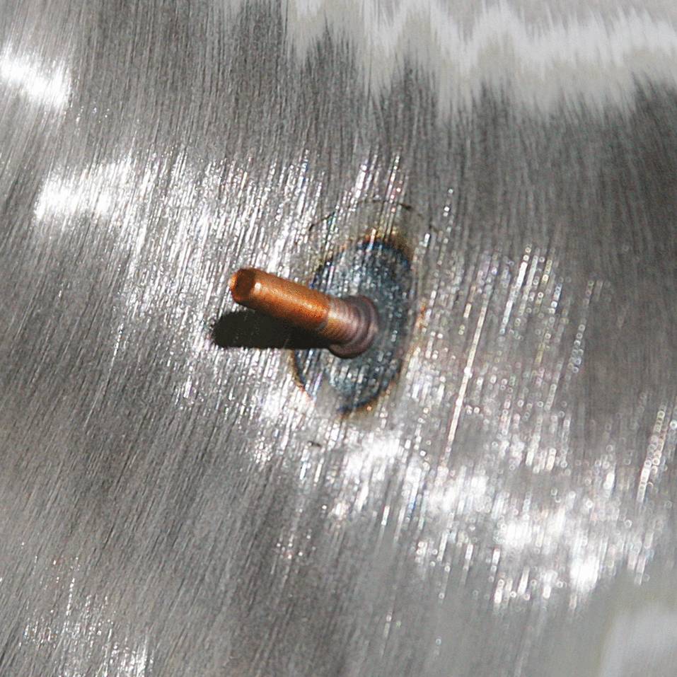 Close-up of a burnished metal pin embedded in a surface. The surrounding area reflects light and shows scratch marks.
