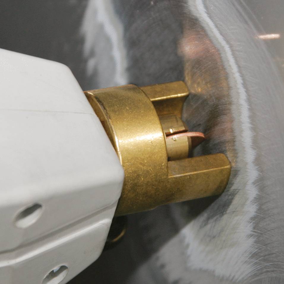 A close-up of a MIG welding torch working at the edge of a metal surface shows a bright arc and sparks.