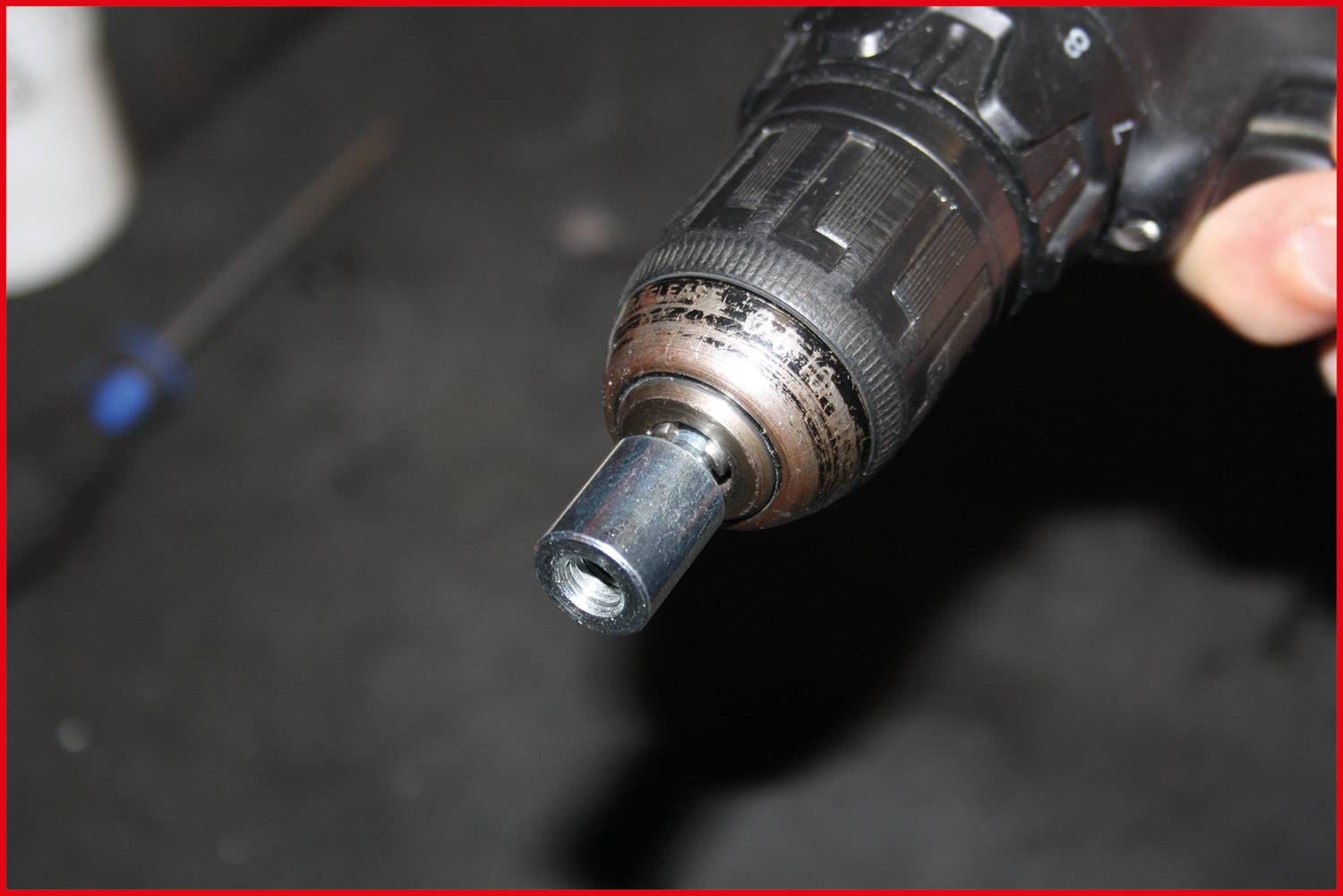 'Cordless drill with attached accessory is being held in hand. A screwdriver can be seen blurred in the background.'