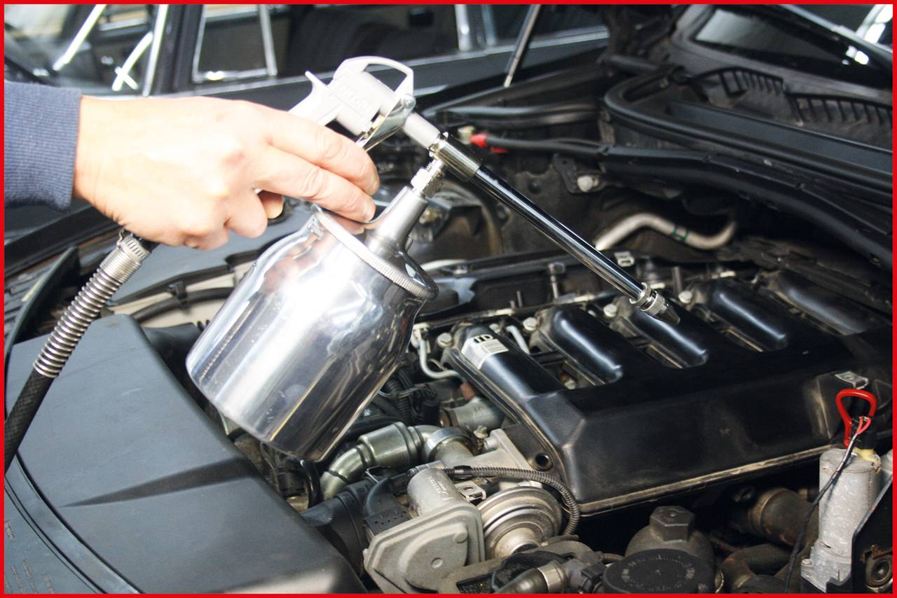 A person is holding a spray gun over an open area of a car, possibly for maintenance or engine cleaning.