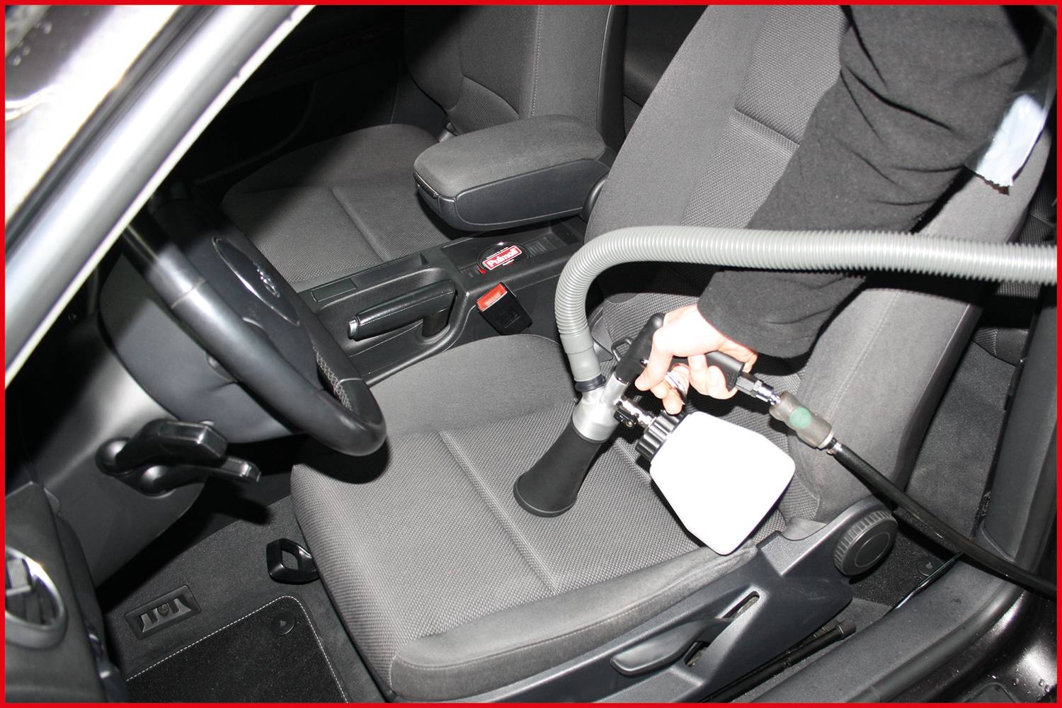A person is cleaning the driver's seat of a car with a vacuum cleaner. A flexible hose is attached to the vacuum cleaner.