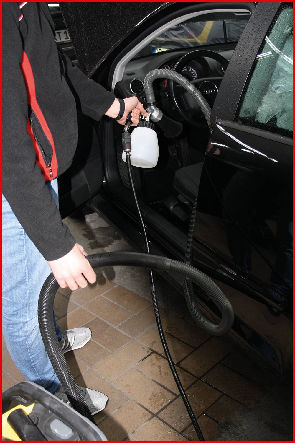 A person is cleaning the interior of a car with professional cleaning equipment. An open car door and a concrete floor are visible.