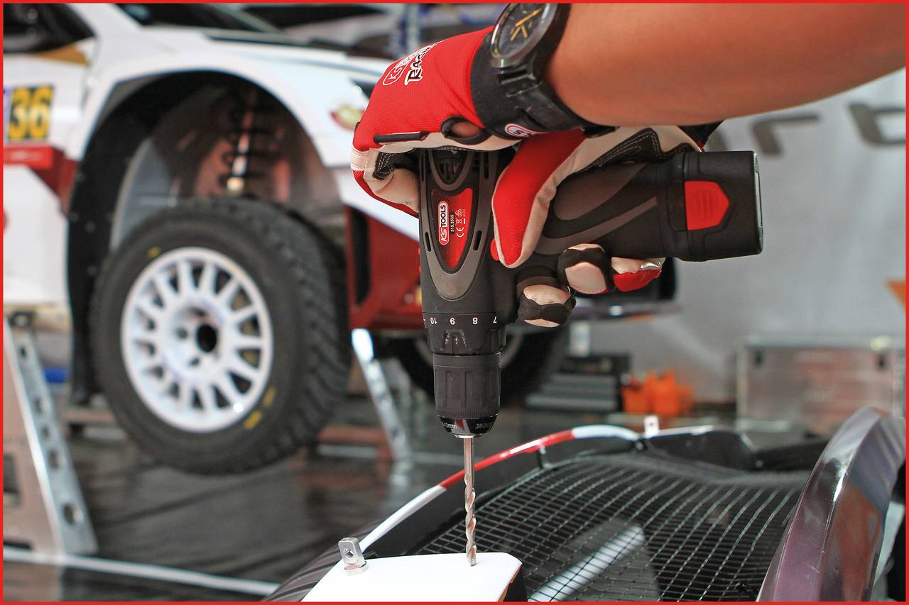 A hand is holding an electric drill over a racing car, with another vehicle jacked up in the background.