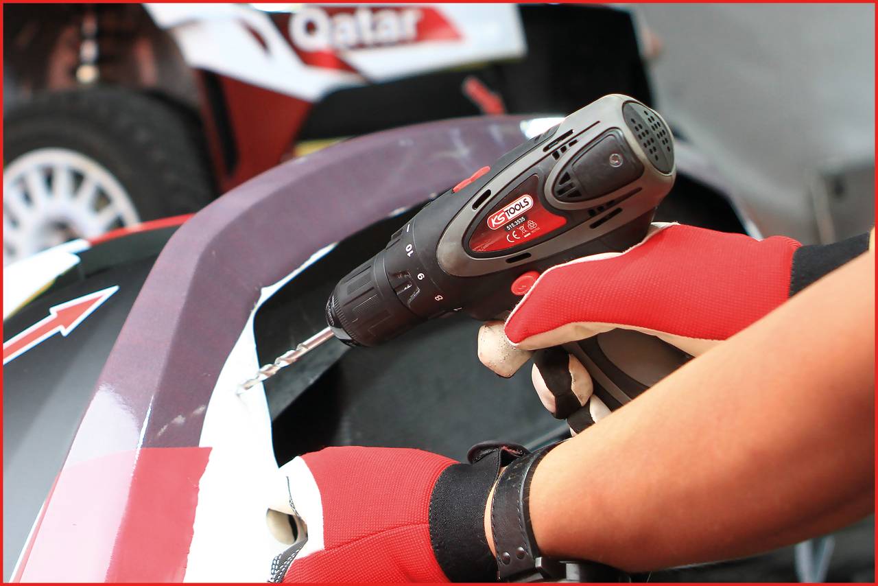 A person is working with a cordless drill on a vehicle part. Car tyres are visible in the background.