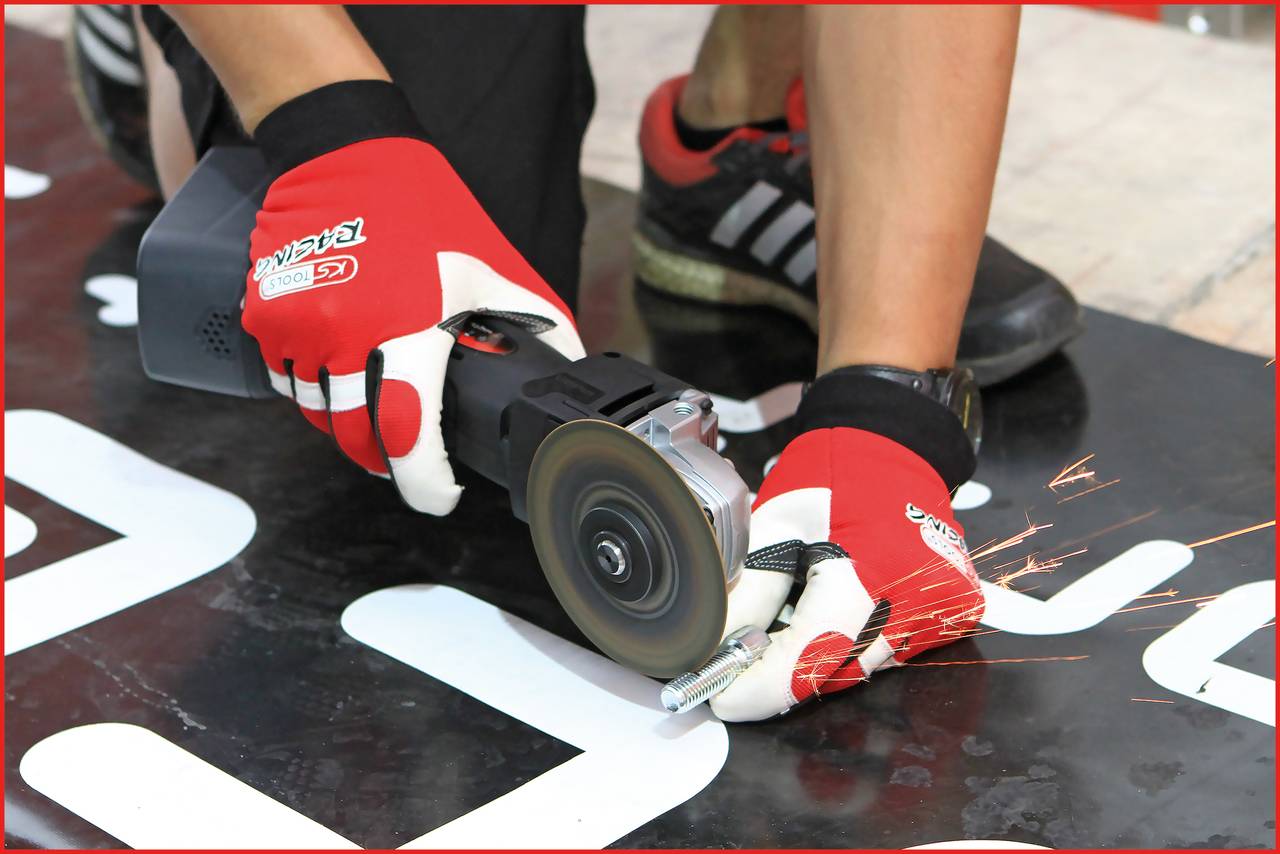 A person wearing red gloves is cutting metal with an angle grinder, with sparks flying. Sports trainers are visible.