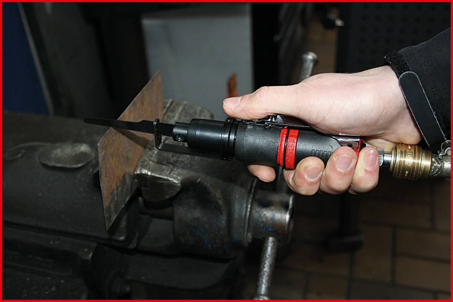 A hand holds an electric tool cutting a metal sheet that is clamped in a vice.