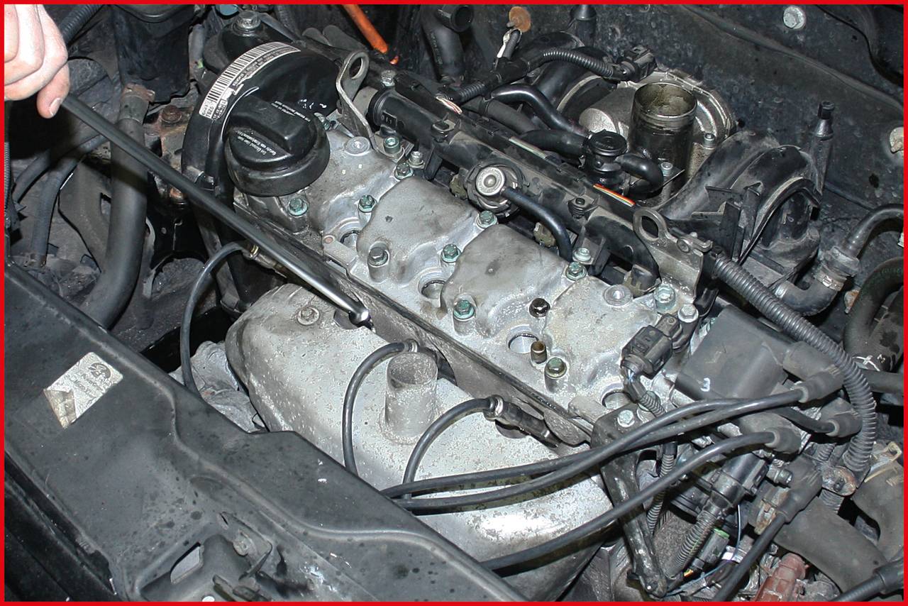 Close-up of a car engine, with visible spark plug surfaces and spark plug leads, being serviced. A hand is holding a tool.