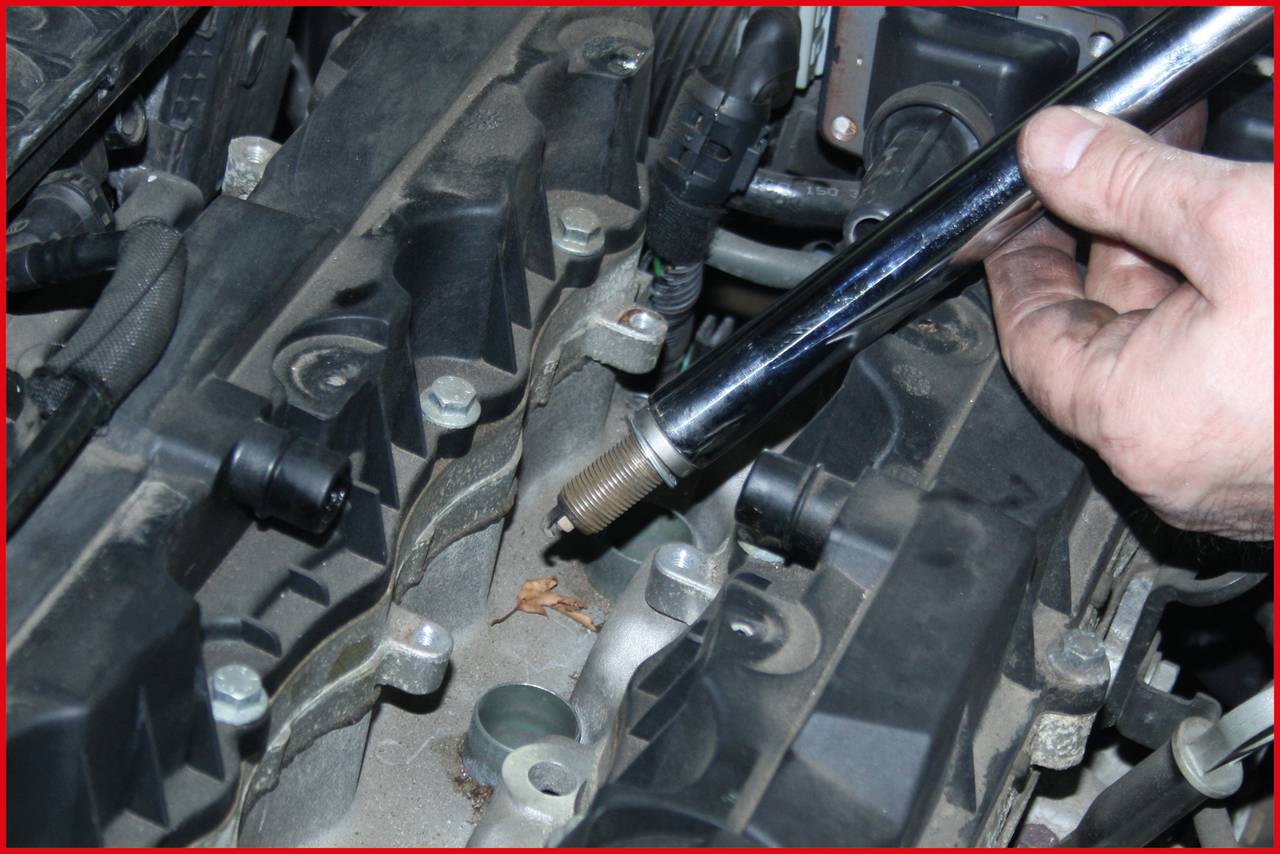 A hand is holding a spark plug over an engine block, ready for installation. Various cables and screws are visible.