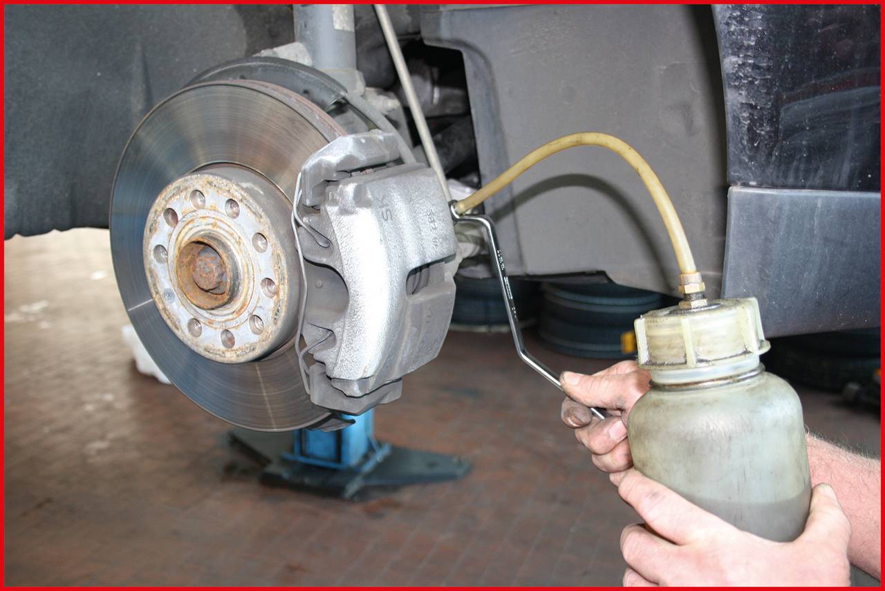 A person is bleeding the brakes of a car. A container is connected via a hose to the brake calliper.