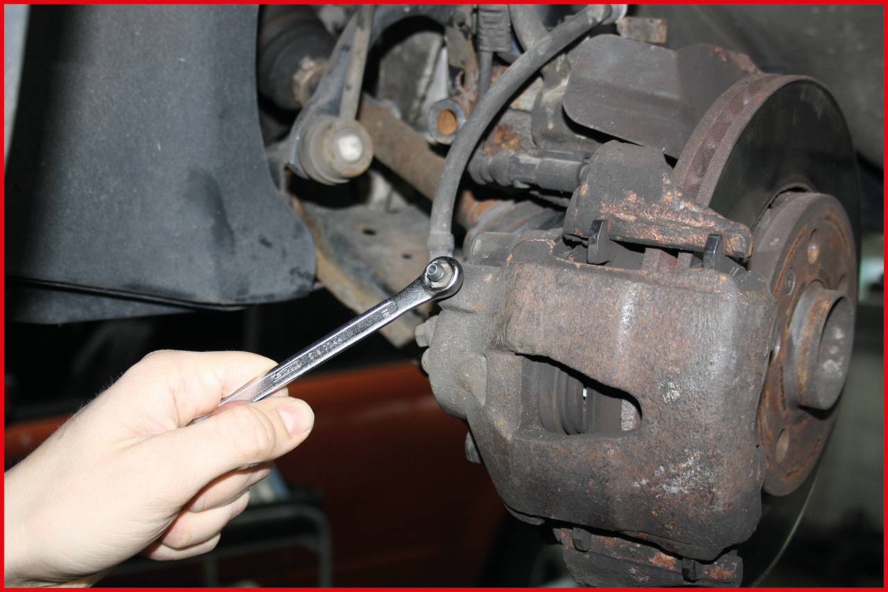 'A hand is holding a spanner and tightening a screw on a vehicle brake system. Vehicle parts are visible.'