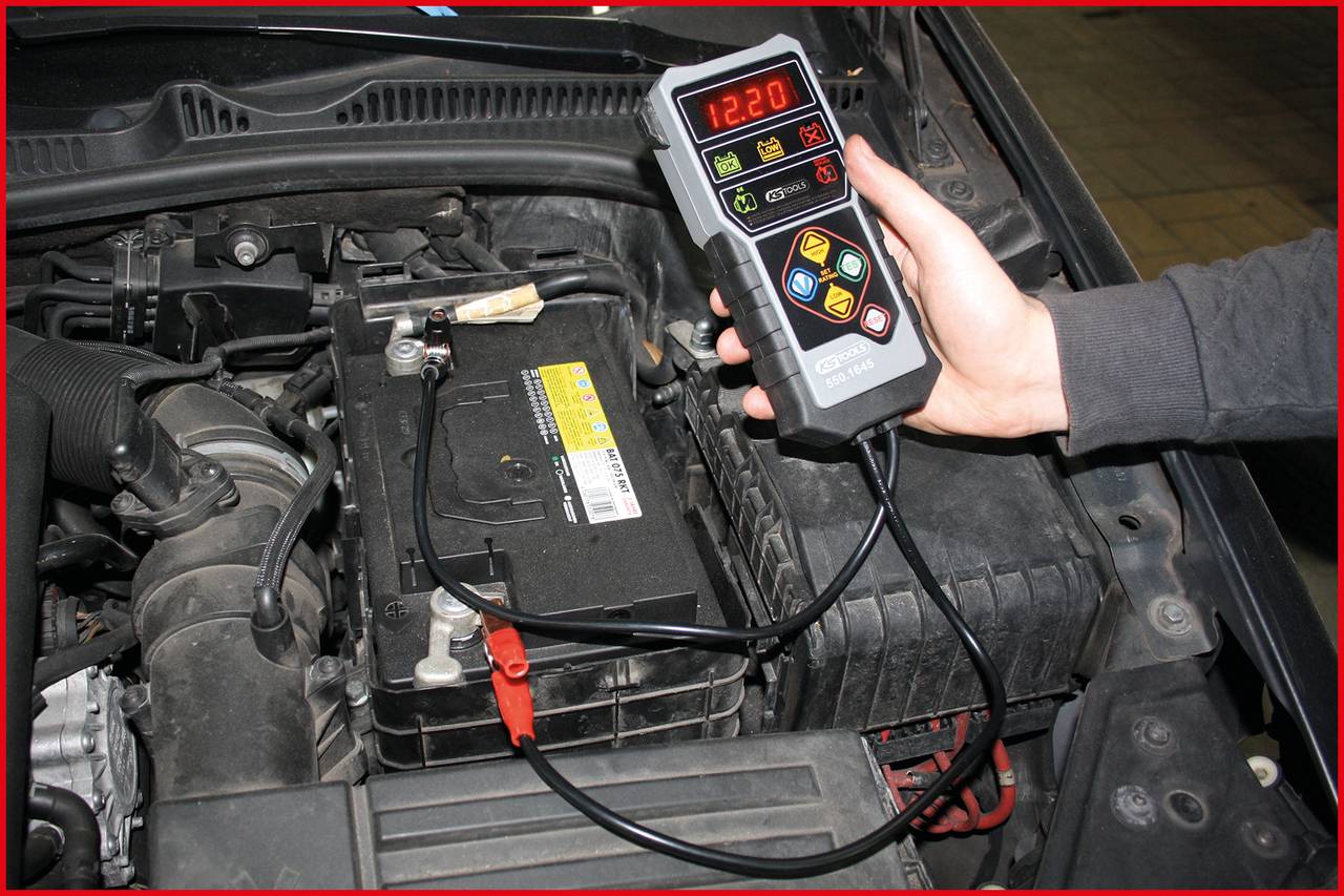 A vehicle technician uses a digital battery testing device to check the car battery. The display shows '12.20'.