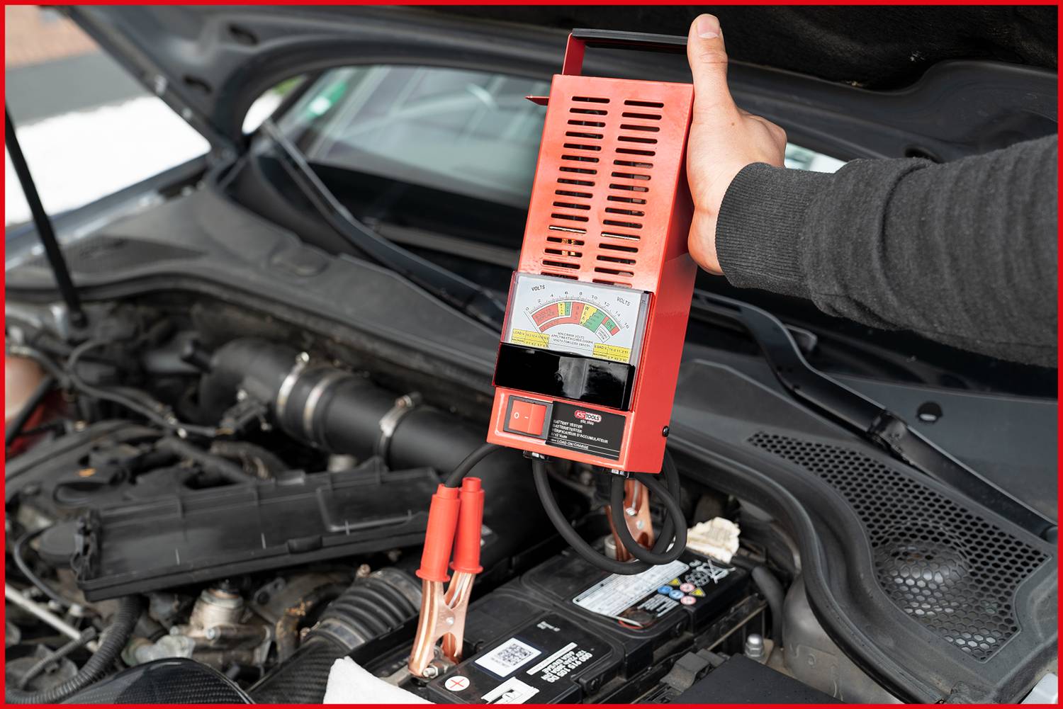 A person is testing the car battery in an open engine compartment with a battery tester. The device displays the charge level.