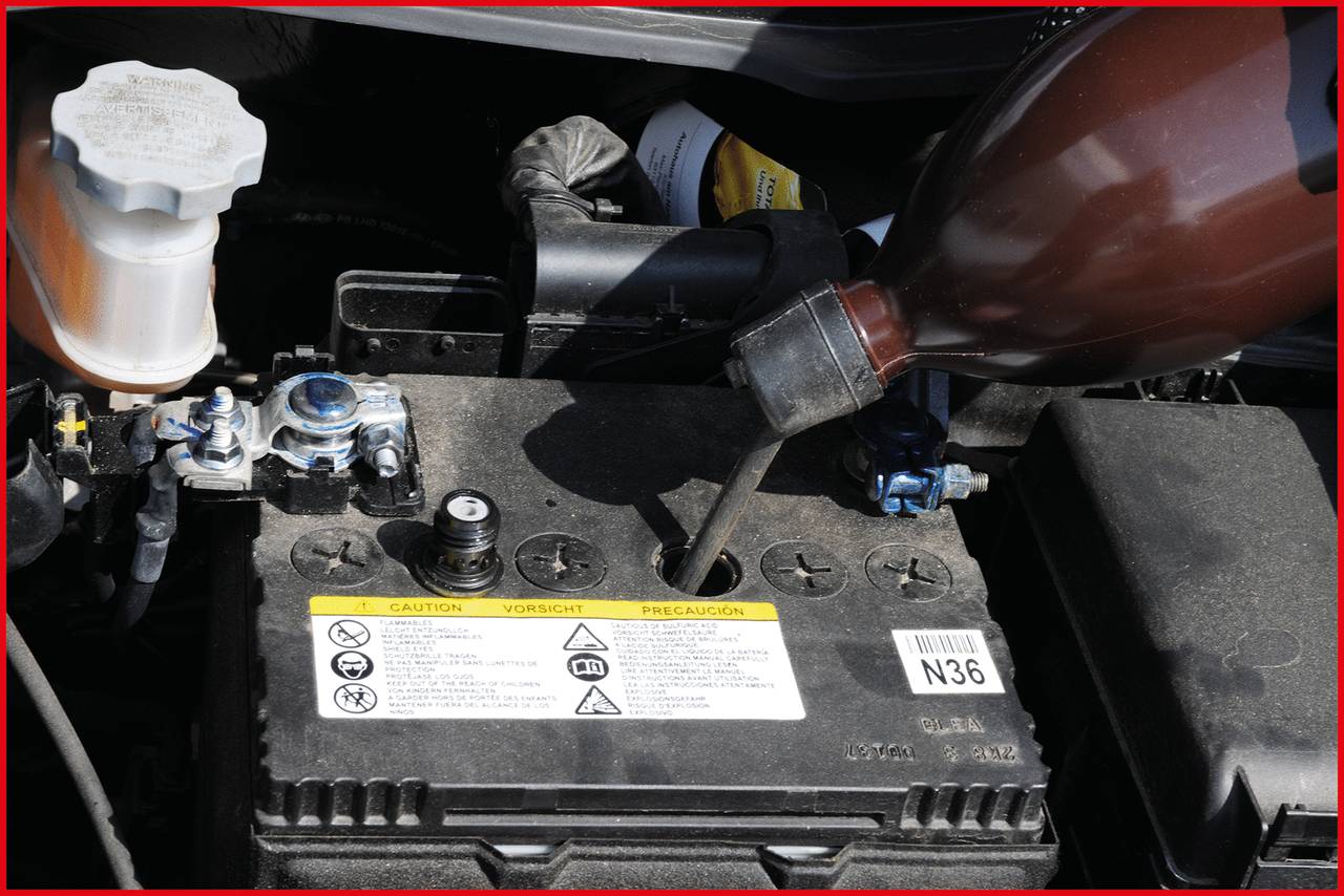 Battery of a vehicle with its lid open, with a plastic bottle nearby topping up the fluid. A container with liquid is visible in the background.