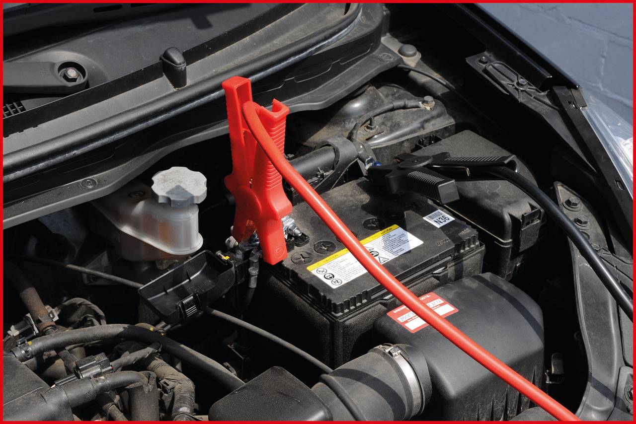 Jump leads connected to a car battery. Red cable on the positive terminal, black cable on the negative terminal. Engine compartment of a car.