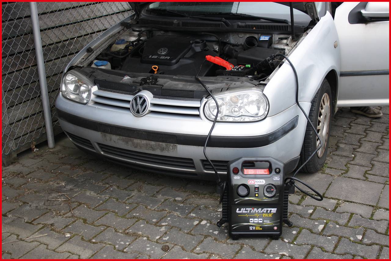A car with an open bonnet is being jump-started using a jump starter. The device is connected to the engine and placed on the ground.