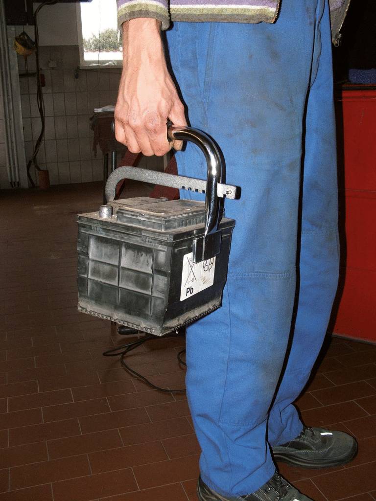 A person wearing blue workwear is carrying a car battery by its handle in a workshop environment.