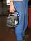 A person wearing blue workwear is carrying a car battery by its handle in a workshop environment.