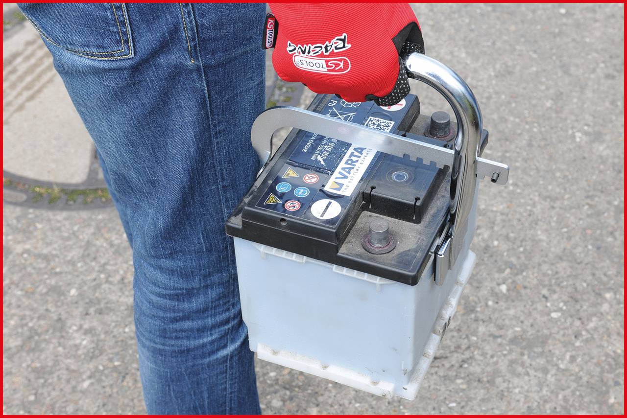A person in blue jeans is holding a car battery with their hand. The person is wearing red work gloves.