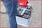 A person in blue jeans is holding a car battery with their hand. The person is wearing red work gloves.
