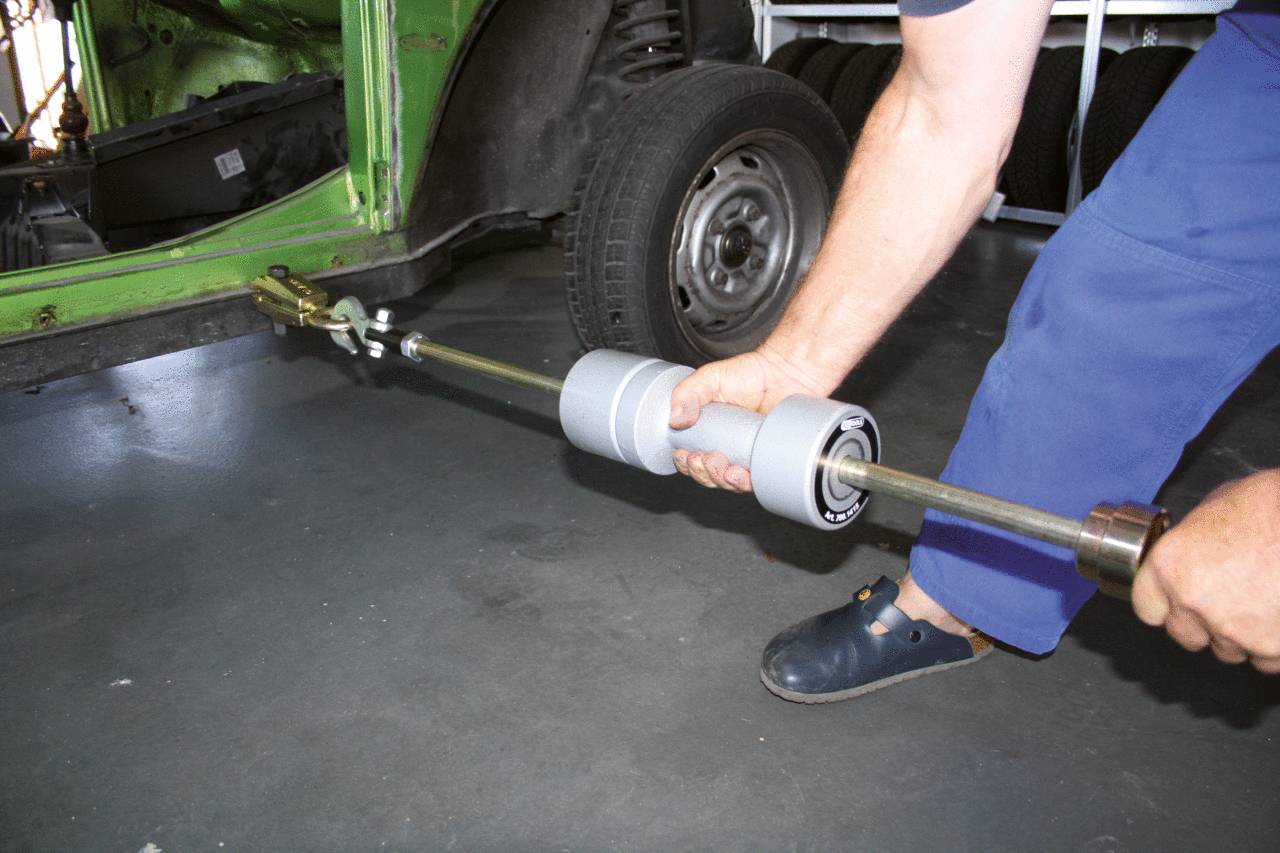 A person is repairing a vehicle frame with a dent removal tool in a car repair workshop. The bodywork is green.