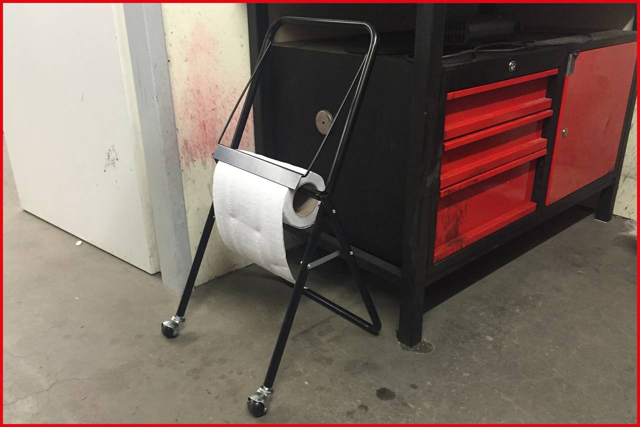 A toilet roll holder on wheels stands in front of a black tool cabinet with red drawers in a workshop environment.