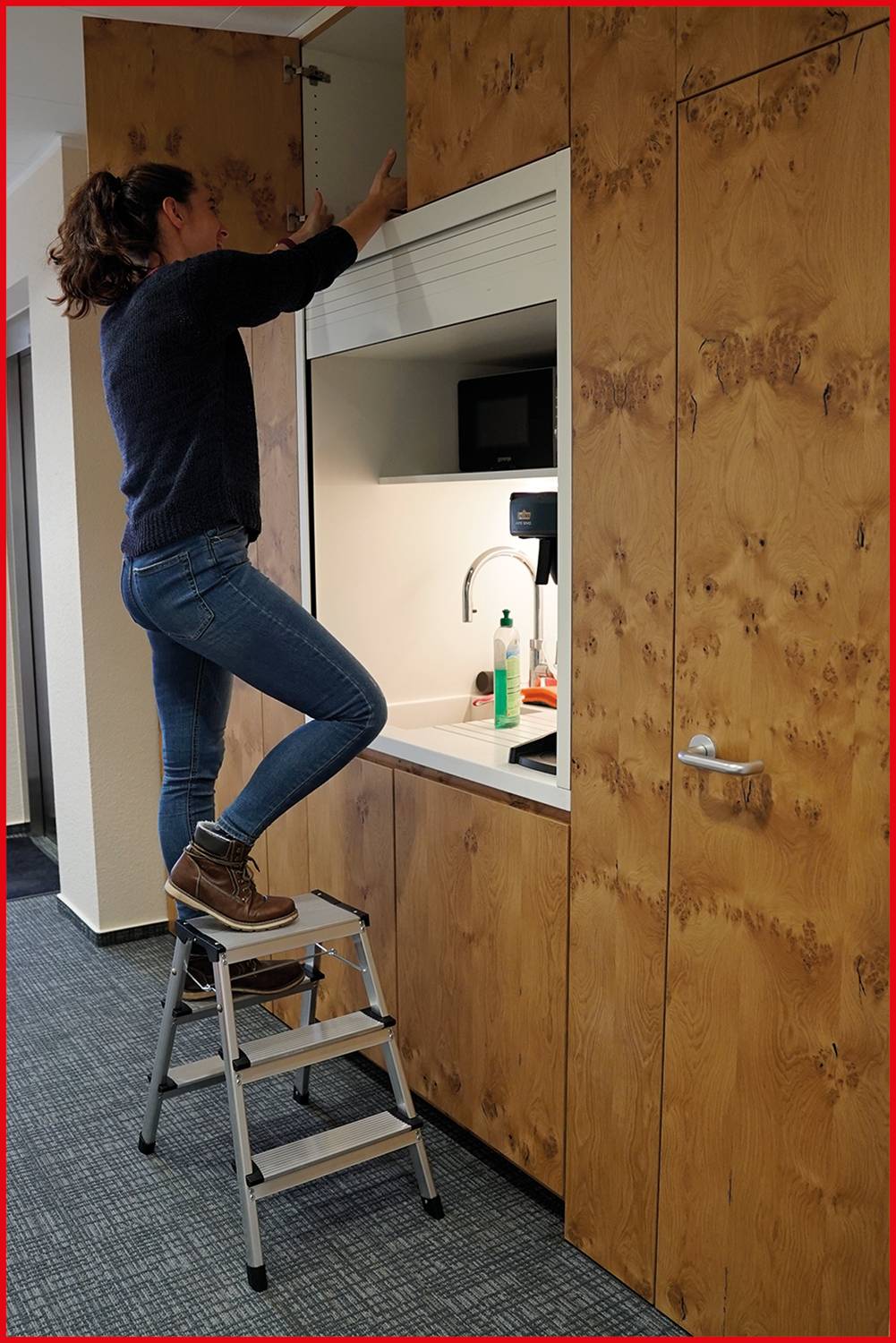 A person is standing on a small stepladder and opening a cabinet above a kitchen unit.