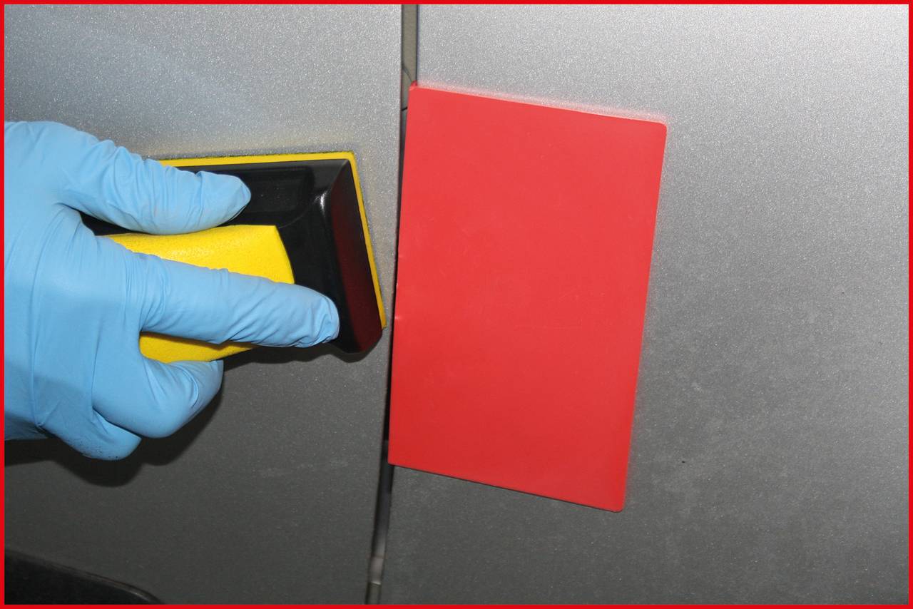 'A hand wearing a blue glove is using a yellow cleaning tool to polish a red rectangle on a grey surface.'