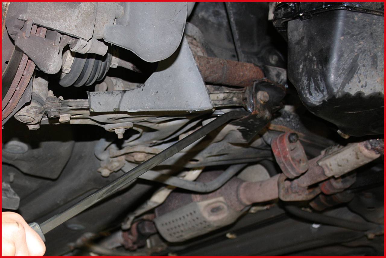 Underside of a car focusing on the exhaust system and suspension. A person is inspecting a component with a tool.