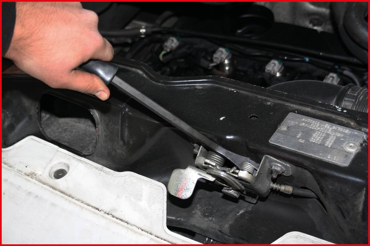 A person pulls a lever under the bonnet of a car. The focus is on engine components and the movement of the lever.