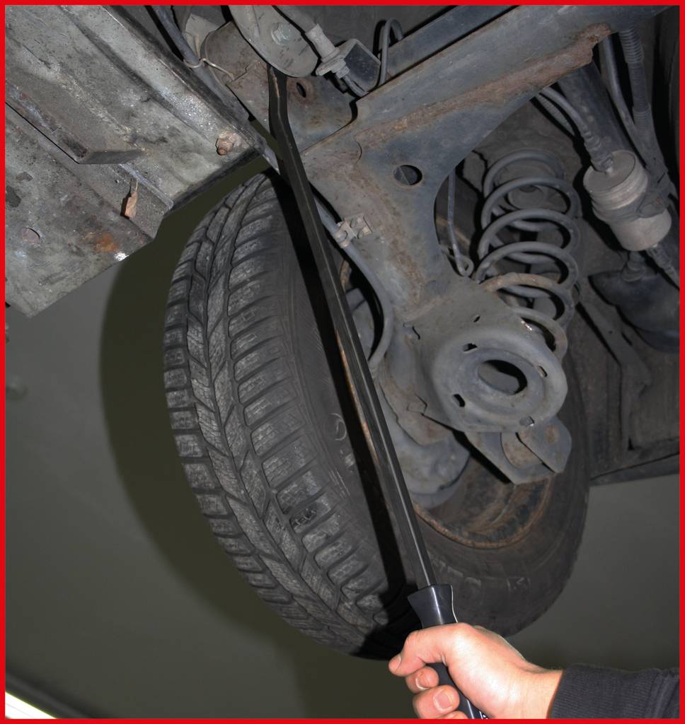 Close-up of a car tyre and suspension, viewed from underneath. A person is holding a tool against the suspension.