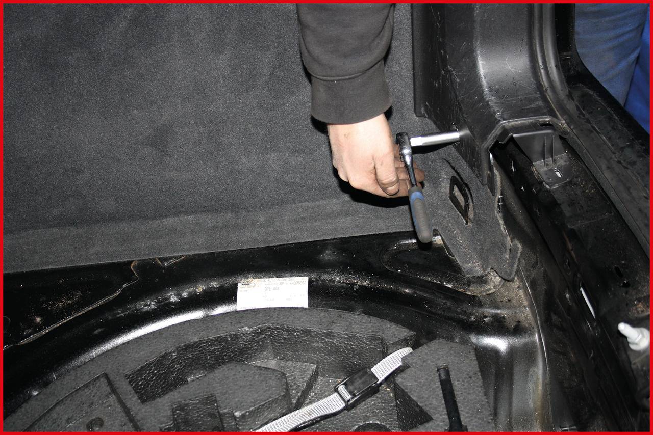A person is tightening a screw in a car boot using a T-bar spanner. A spare tyre is visible.