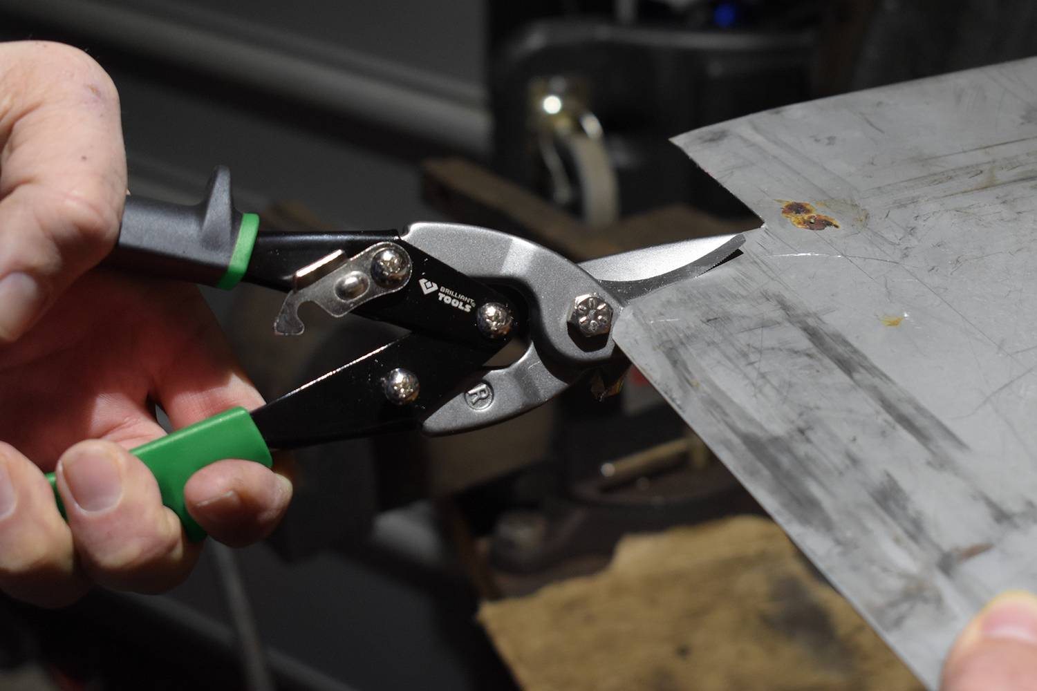 A person is cutting a piece of sheet metal with a green and black handheld tin snips. A workbench is visible in the background.